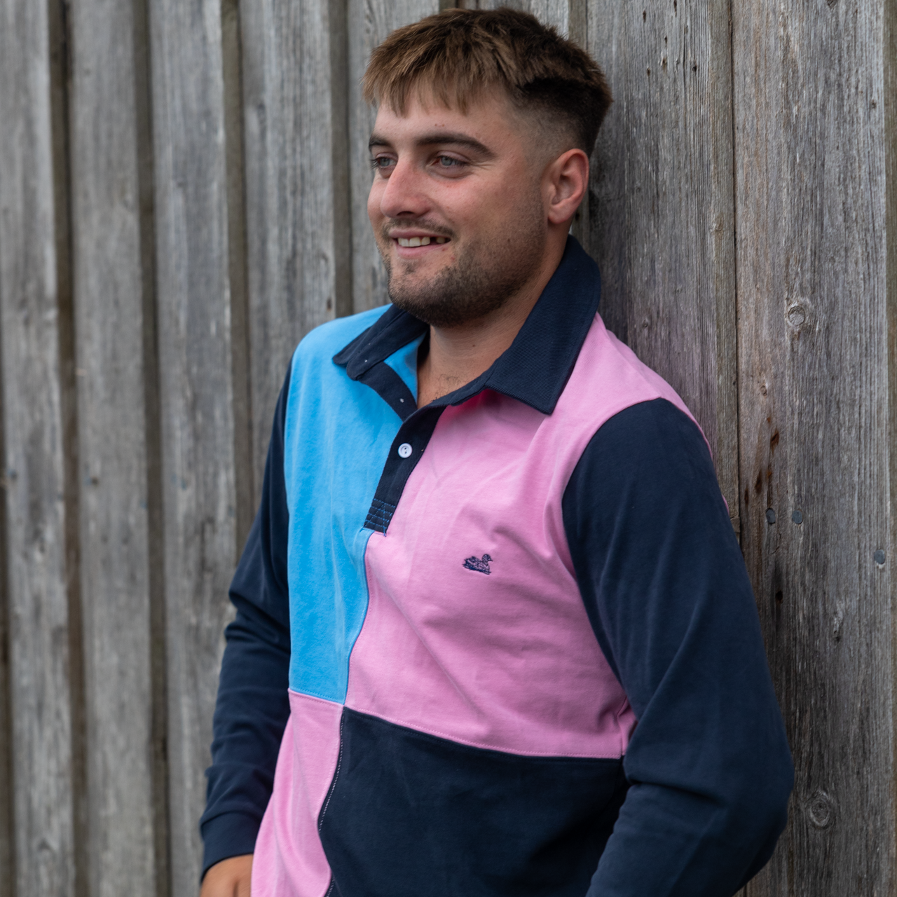 A person wearing a vintage style rugby shirt with color blocks in pink, blue, and navy, standing against a wooden backdrop.